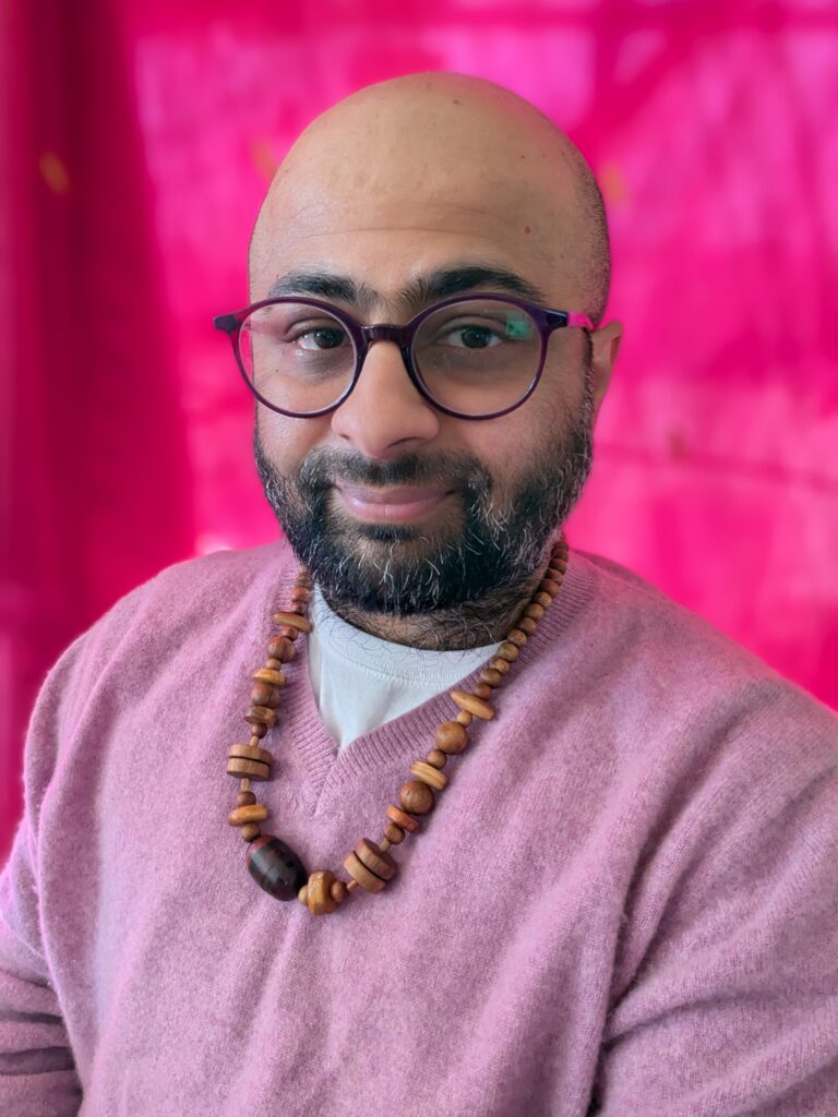 headshot of Anand, a bald brown-skinned man with salt-and-pepper beard cropped close. he wears round purple-rimmed glasses, a lavender sweater, and a wood-bead necklace in front of a pink background. 